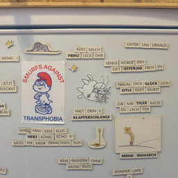 Part of a refrigerator door with stickers and magnetic words from Le Petit Prince. There is a "Smurf's against Transphobia" sticker and a uxn sticker with Varvara as well as magnets with illustrations from the book. The "poems" are in German language and make little sense, especially translated to English. "Küss mich Prinz Leckohr" — "Kiss milk, prince lick ear" or "Adieu Frau Käseklug, Herz König Echo Ei muss nie mehr erwachsen tun." — "Adieu, Ms. Cheesesmarts, King of Hearts Echo Egg doesn't ever have to pretend to be grown up again." or "Kauf wenig!" — "Buy little!"