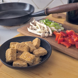 A cast iron pan with a beautiful wooden handle on a stove top. Next to it is a small bowl with breaded tofu cubes. Besides it a wooden board with mushrooms, cut bell peppers and some chopped spring onions on it.