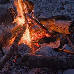 Closeup of a campfire by a small river at early night time. The fire is burning brightly and is circled by a few larger rocks. Between the rocks is an iron cast pan containing a filled tortilla.