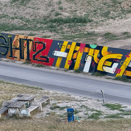 View from a bridge onto a painted construction fence showing the letters AIGHTZ and HITZE.