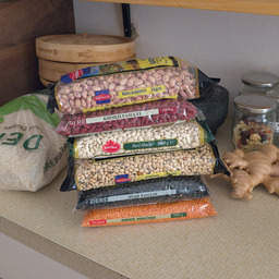 Six bags worth ~5kg of differently shaped, sized and colored beans on a kitchen counter. Next to it is a 5kg bag of rice as well as a fairly large piece of ginger root.