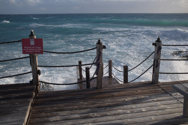 Waves crashing against the dock with some wooden stairs leading into the water. There is a sign that reads "Betreten auf eigene Gefahr" ("Enter at your own risk").