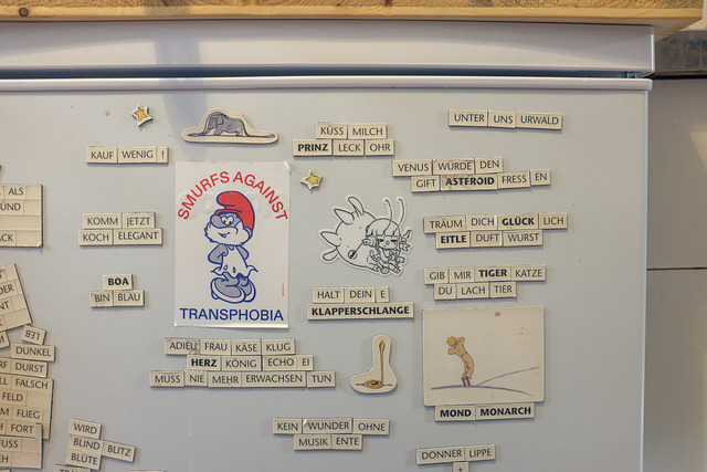 Part of a refrigerator door with stickers and magnetic words from Le Petit Prince. There is a "Smurf's against Transphobia" sticker and a uxn sticker with Varvara as well as magnets with illustrations from the book. The "poems" are in German language and make little sense, especially translated to English. "Küss mich Prinz Leckohr" — "Kiss milk, prince lick ear" or "Adieu Frau Käseklug, Herz König Echo Ei muss nie mehr erwachsen tun." — "Adieu, Ms. Cheesesmarts, King of Hearts Echo Egg doesn't ever have to pretend to be grown up again." or "Kauf wenig!" — "Buy little!"