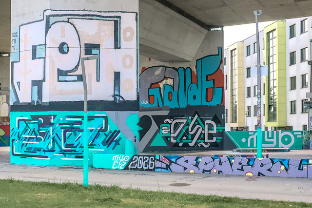 A mural combined over two large concrete pillars. One is a lot further away than the other, but from the right perspective both pieces merge into one. On the left, there are abstract letters spelling EIZ, on the right, it spells @EASE. In the center there is a real and black colored broken heart.