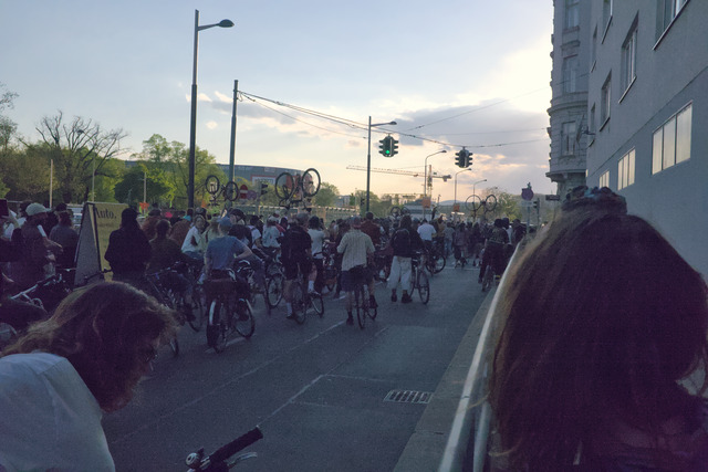 A city road filled with people on bikes, currently idling. Some of them lift their bikes up in the air.