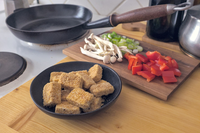 A cast iron pan with a beautiful wooden handle on a stove top. Next to it is a small bowl with breaded tofu cubes. Besides it a wooden board with mushrooms, cut bell peppers and some chopped spring onions on it.