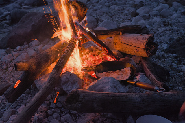 Closeup of a campfire by a small river at early night time. The fire is burning brightly and is circled by a few larger rocks. Between the rocks is an iron cast pan containing a filled tortilla.
