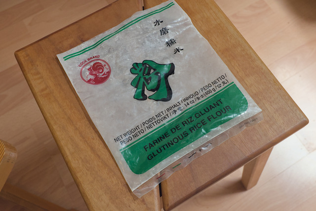 An empty bag of rice flour on a wooden stool. It has a red rooster as a logo on it that proudly states "Cock Brand" on top of it.