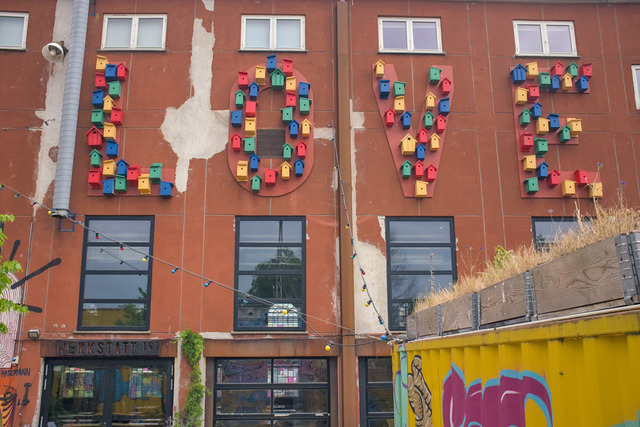 A rusty metal plated wall that carries large cut out letters spelling LOVE. The space of each letter is filled with differently colored and shaped bird houses.