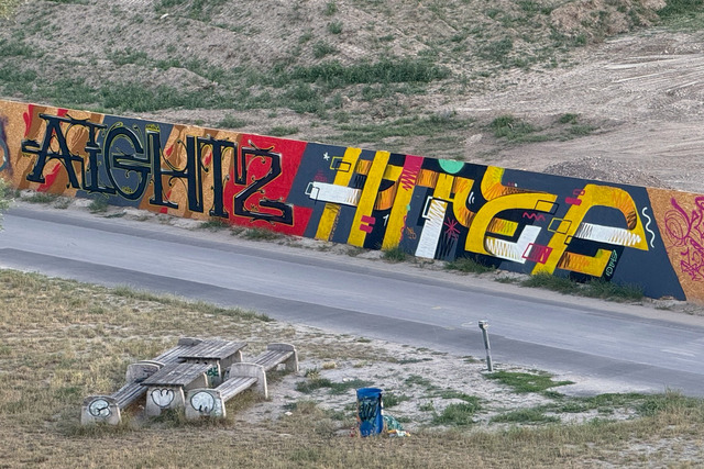 View from a bridge onto a painted construction fence showing the letters AIGHTZ and HITZE.