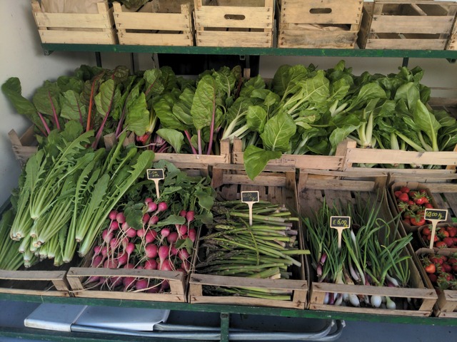 View into a a trailer opened towards the sides. It contains a few shelves filled with wooden boxes containing spring onions, ruccola, mangold, spinach, asparagus and strawberries. Everything is nicely arranged and looks fresh and colorful.